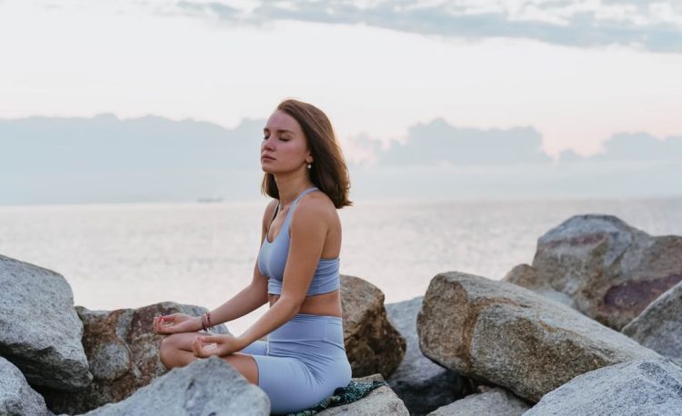 Woman in activewear meditating peacefully on rocks by the ocean at sunrise, promoting wellness and tranquility.