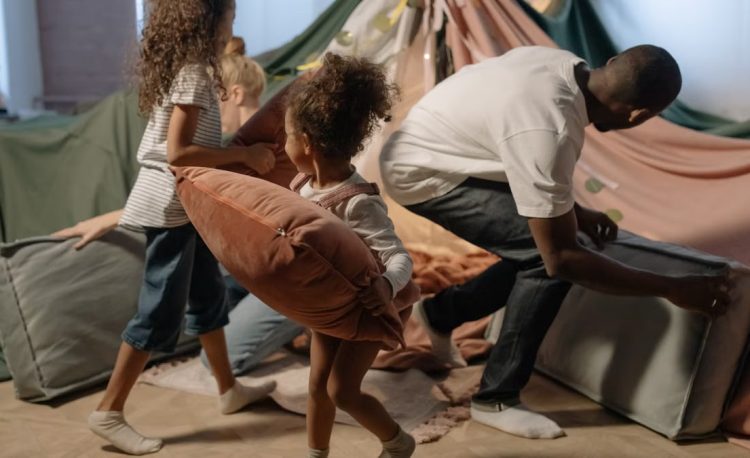 A playful family scene with a father and daughters having a pillow fight inside a cozy indoor fort.